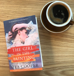 The Girl in the Painting by Tea Cooper is pictured on a wooden table next to a cup of black goffee